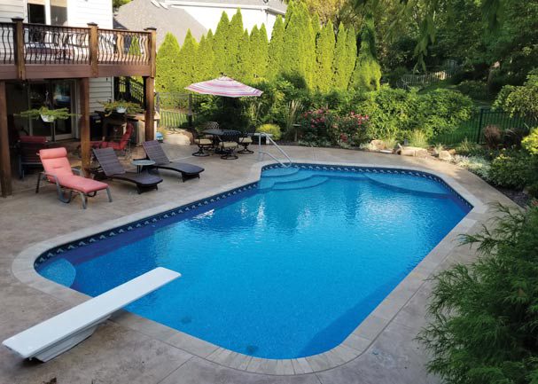 Latham-Vinyl-Pool-Example-Azure-Pools Rectangular backyard swimming pool with a diving board, lounge chairs, patio tables, and surrounding greenery on a sunny day—perfect for comparing fiberglass vs. concrete vs. vinyl pool options.