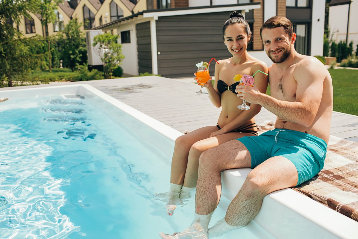 azure-fiberglass-oll–lovely-heteroseksual-couple-rented-a-house-with-a-2025-09-14-15-45-01-utc A man and woman sit on the edge of a backyard fiberglass pool with their feet in the water, holding colorful drinks and smiling at the camera.